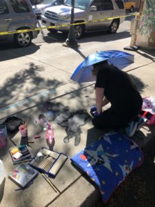 Maya, a teenage girl, drawing on the sidewalk for the Pastels on Fifth event. She is wearing an umbrella hat to stay cool.