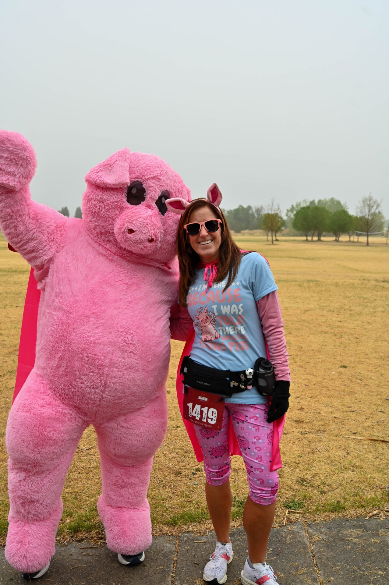A person in a pink pig costume stands next to a smiling woman wearing pink pig-themed clothes, sunglasses, and a race bib numbered 1419. They pose together outdoors on a grassy area with trees in the background.