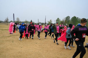 A group of people wearing pink shirts and jackets participate in an outdoor charity run or walk event on a cloudy day, moving across a grassy field with some trees in the background.