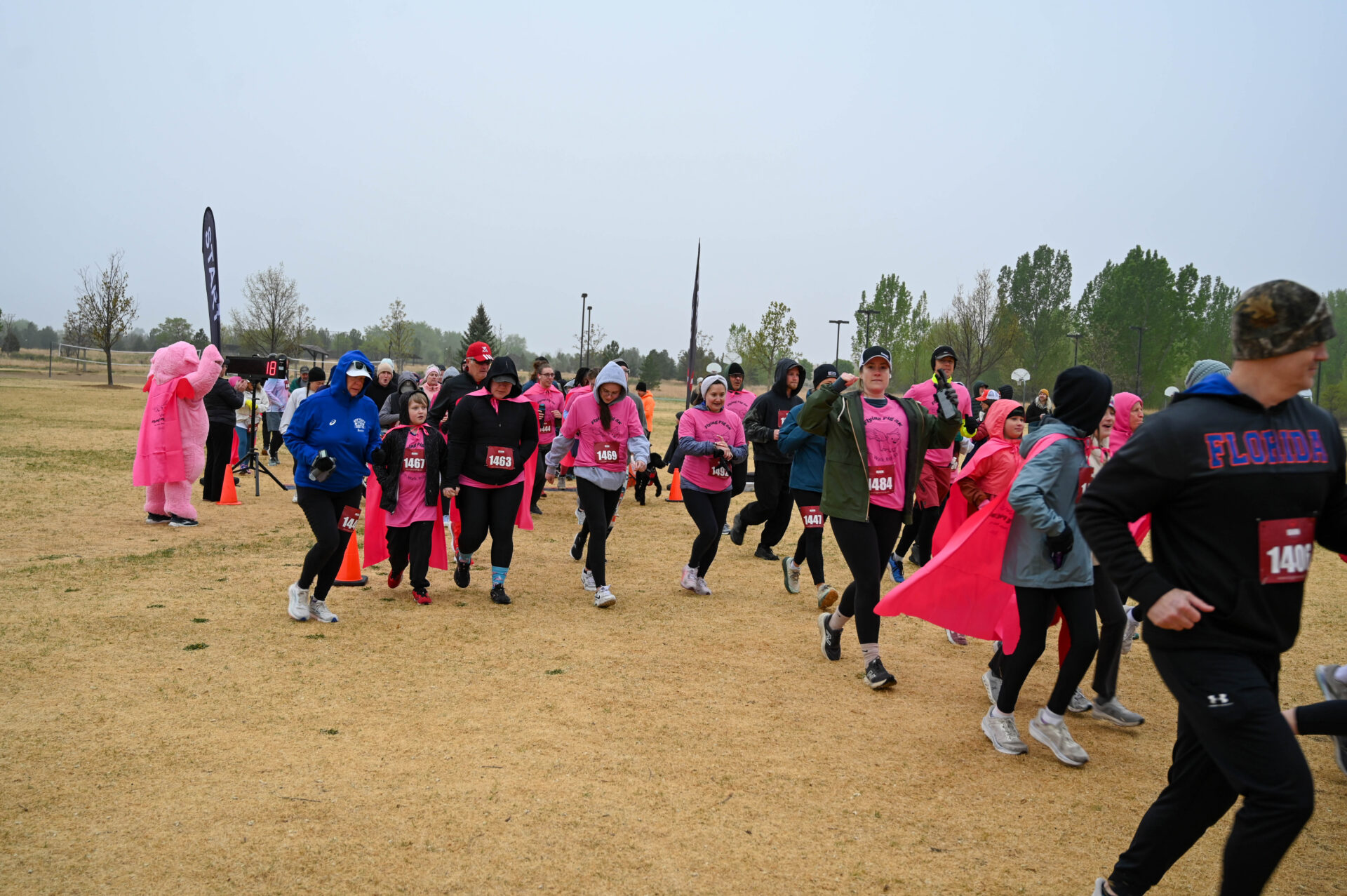 A group of people wearing pink shirts and jackets participate in an outdoor charity run or walk event on a cloudy day, moving across a grassy field with some trees in the background.