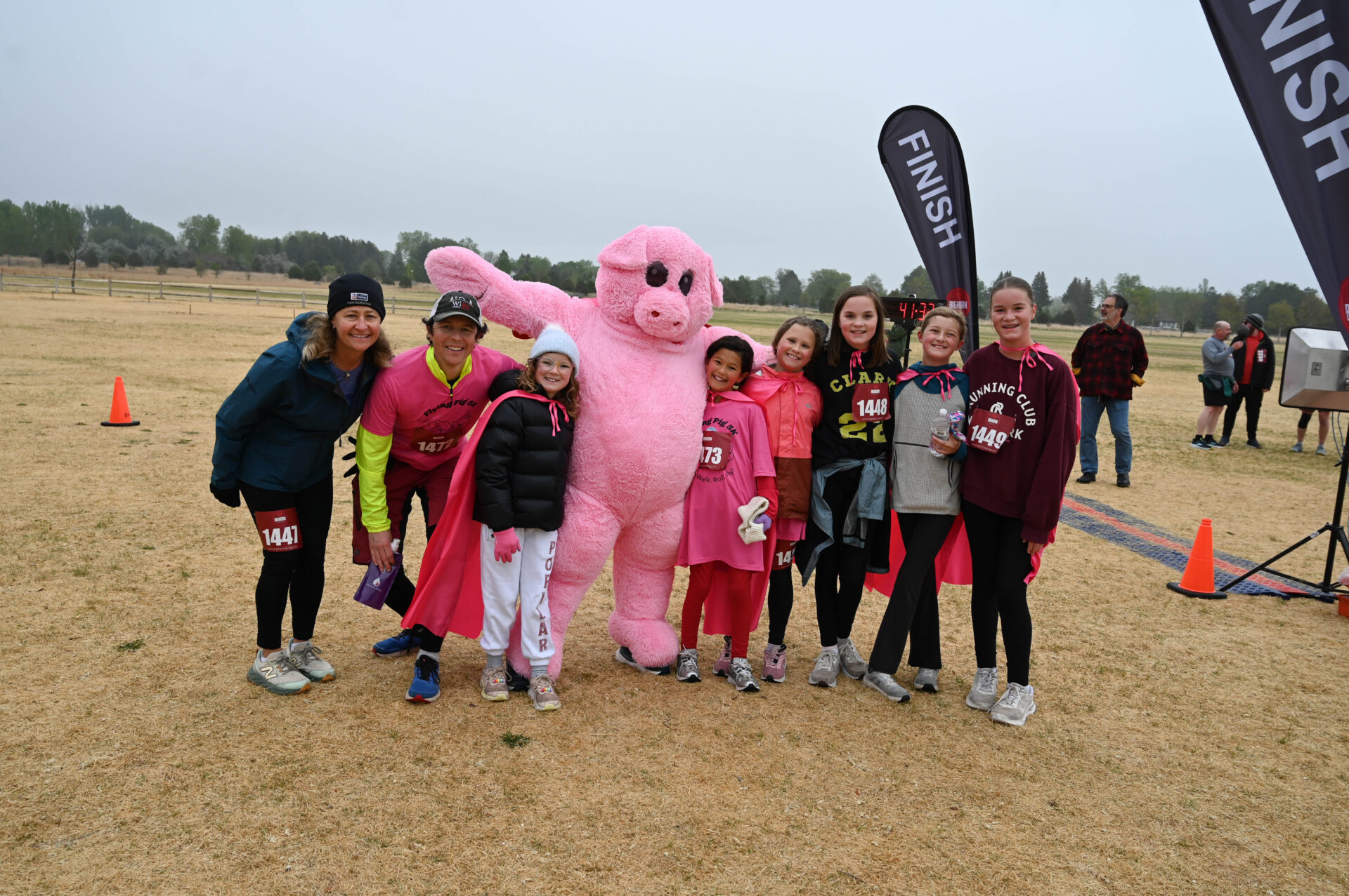 A group of smiling people, some wearing race bibs and pink capes, stand together outdoors next to a person in a pink pig costume near a finish line banner on a grassy field.
