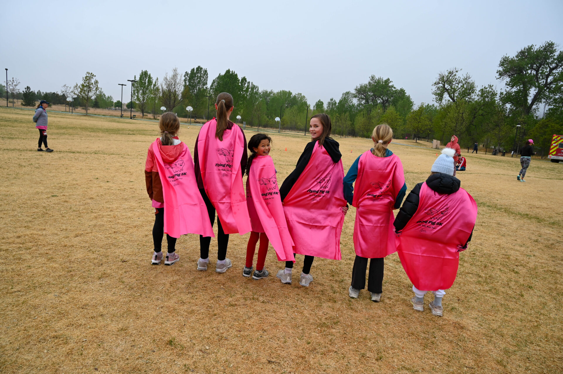 Six children stand in a line outdoors on dry grass, facing away from the camera. They wear bright pink capes, and trees and more people are visible in the background under a cloudy sky.