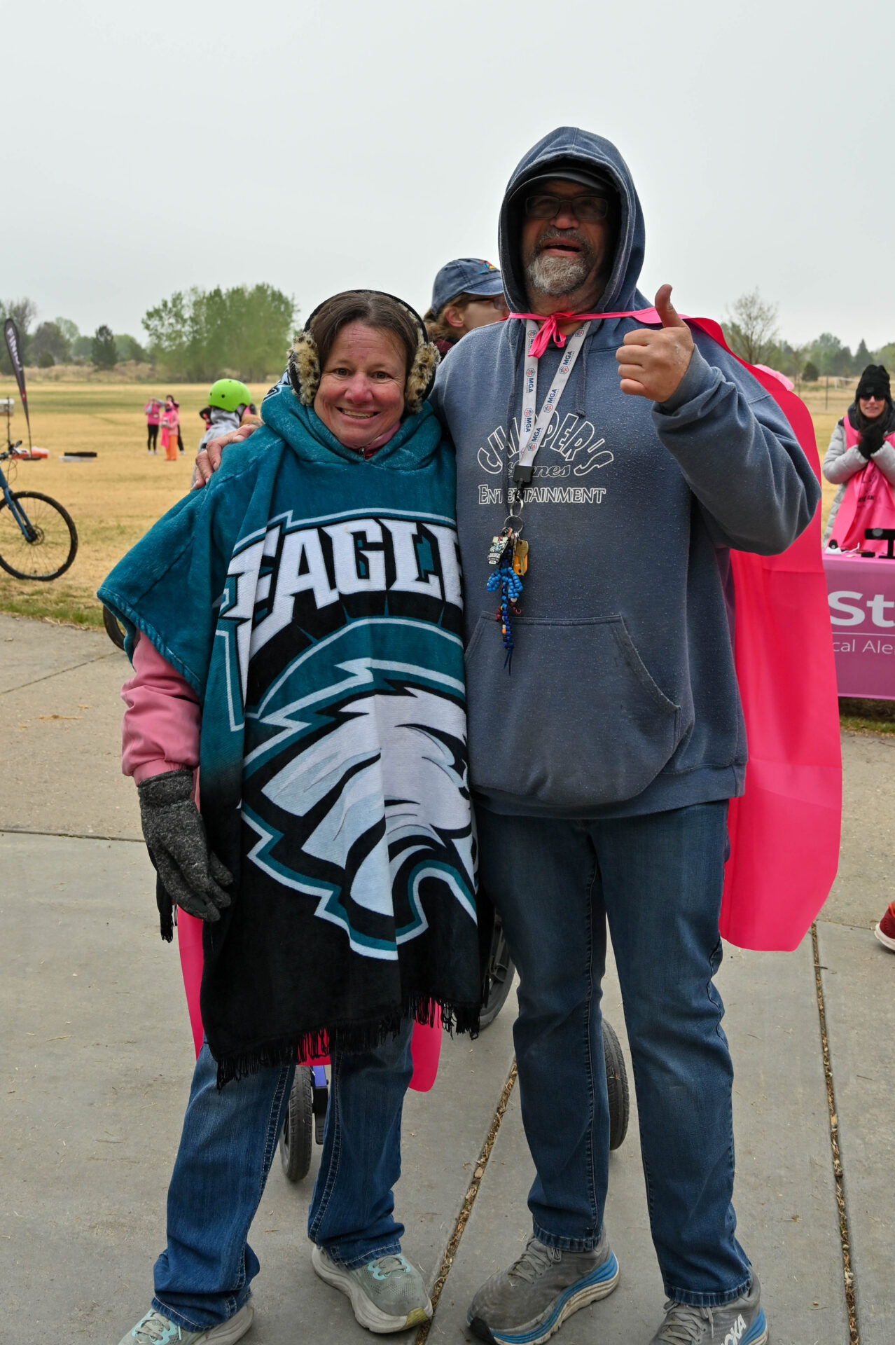 Two people bundled in warm clothes and hoodies stand together outdoors, smiling. One is wrapped in an Eagles blanket, and the other gives a thumbs up. Pink event capes and participants are visible in the background.